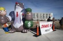 Three men can be seen standing outdoors on a corner of an intersection. They are standing behind a banner that reads "Shame on Port KC." At left is a large inflatable cat that is is smoking a cigar wearing a suite. It  has its paws wrapped around an inflatable person who looks like a construction worker in its right paw and a bag of money in its left paw.