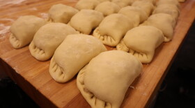 A 4x4 row of pasties sits on a large wooden table. The half-moon shaped pastries are crimped along the outer edge. The dough is still a light cream color. After they're baked, they'll be a golden brown!