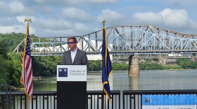 Andy Beshear stands behind a podium wearing a suit coat and sunglasses. Behind him is the Ohio River and several bridges in the distance.