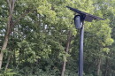 A black license plate reader and solar panel sit on the top of a pole, with woods in the background.