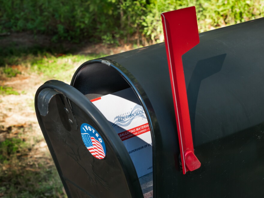 A black mailbox with an "I voted" sticker on it, slightly ajar. There is a mail-in ballot inside and the red mailbox flag is up.