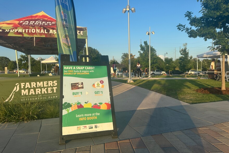 A sign is propped up alongside a farmers market tent in a park.