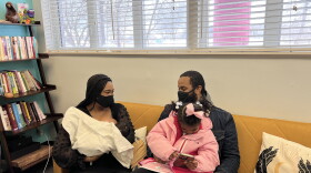 Novia Williams-Later is sitting on a yellow couch next to her husband Deontre. She is holding her newborn baby Ahmir. Ahmir is bundled up in a white blanket. Nova is wearing a black dress and a mask. Deontre is holding the couple's daughter Aria. He is wearing a mask and a jean jacket. Aria is four years old and is wearing a pink jacket and a pink ribbon on the top of her head. Her hair is braided. The Williams-Lasters are African Americans with medium brown skin. 