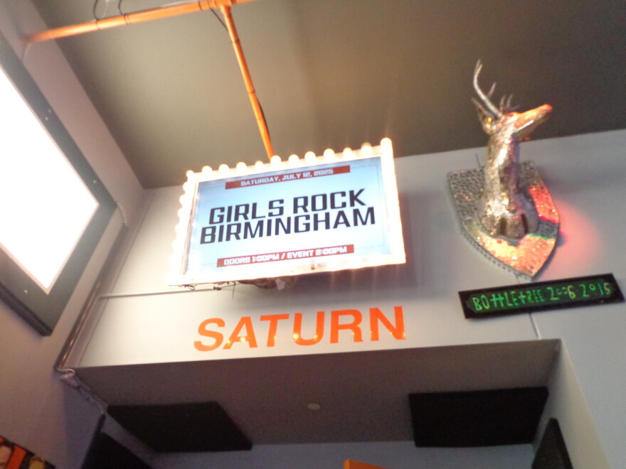 An indoor sign under marquee lights displaying "Girls Rock Birmingham" above a "Saturn" wall decal.