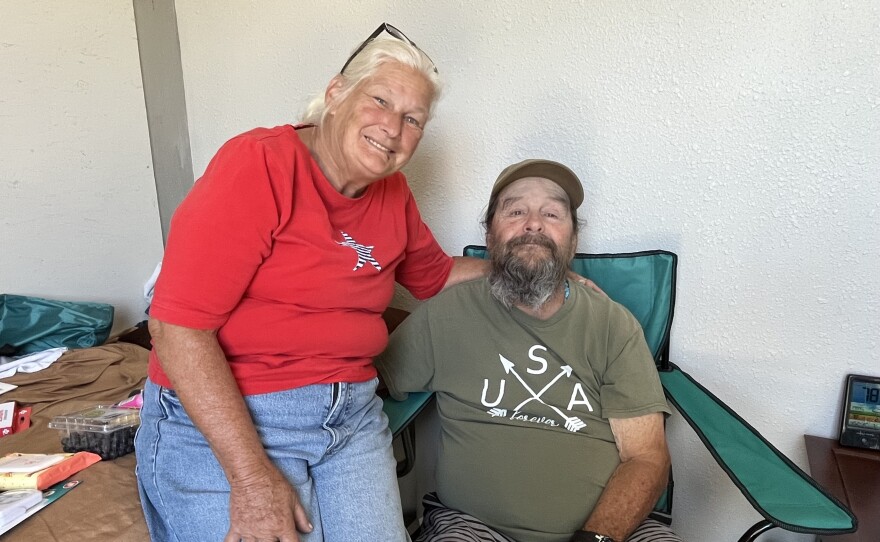 A middle-aged man and woman sit with their arms around each other. She has white hair and he has a long gray beard. 