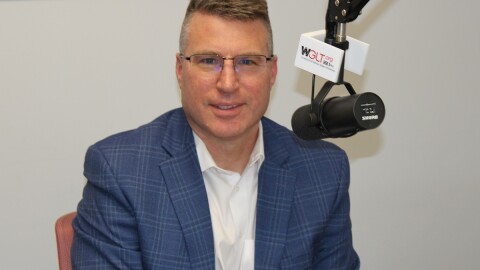 A man in a suit jacket, dress shirt and eye-glasses sits in a chair with a microphone in front of him. 