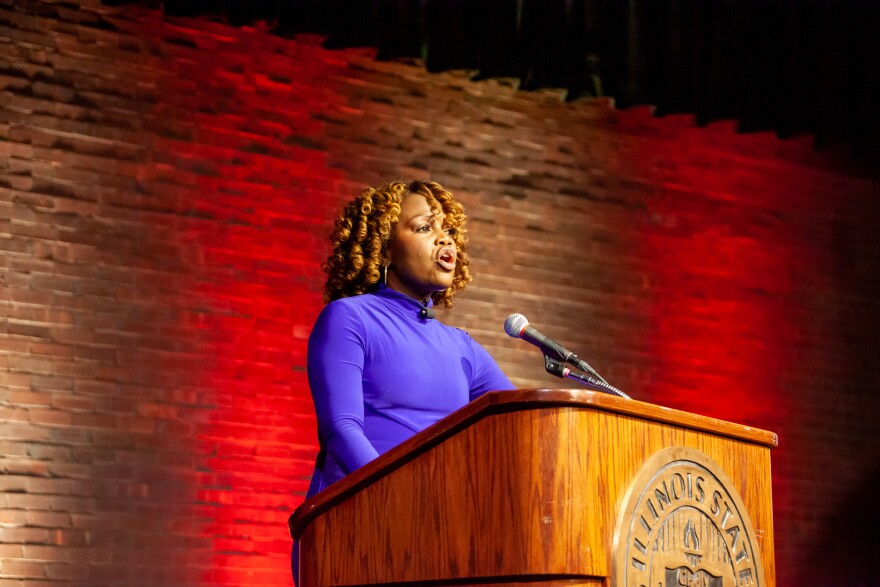 Karine Jean-Pierre stands at a wooden podium delivering a speech. She is wearing a blue dress, hoop earrings and curled hair. 