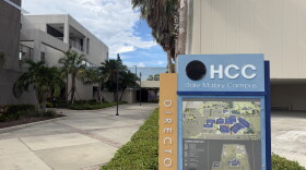 A blue and tan directory sign with a map of Hillsborough College's Dale Mabry campus stands in front of the entrance of grey classroom buildings and trees lining the walkway.