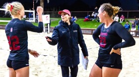 a beach volleyball coach in a black nike jacket and long pants carrying a clipboard talks to two players in black tops with red numbers and b lack shorts on a sandy beach volleyball court