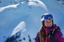 A woman wearing a ski helmet, goggles, jacket and backpack smiles. Behind her is a massive snowy peak dazzled by sunlight.