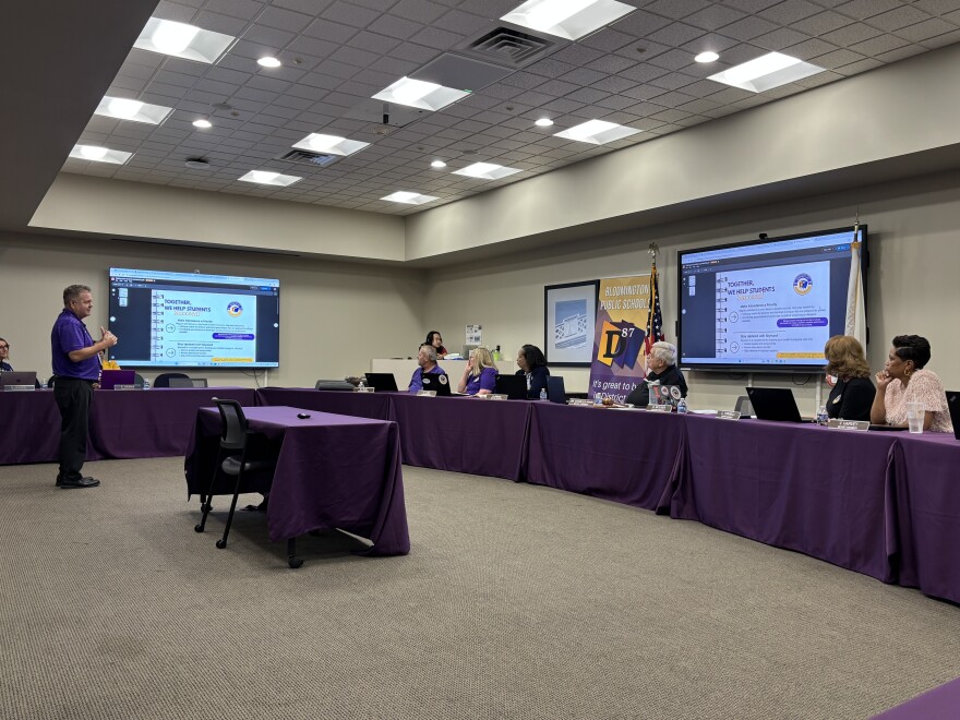 A man stands in front of a group assembled around a board room, presenting on a large screen. 