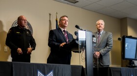 Three men stand in front of a podium at a press conference. A man in a black suit and burgundy striped tie, Tim O'Hare, is speaking. To his left stands Bill Waybourn, dressed in his black sheriff's uniform. Phil Sorrells stands to his right in a gray suit, smiling slightly.