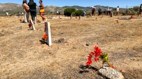 Un campo despejado de arbustos es el hogar de muchas tumbas, y la mayoría están marcadas con una piedra desgastada. A lo lejos hay gente visitando varias tumbas y un grupo de mujeres hablando.