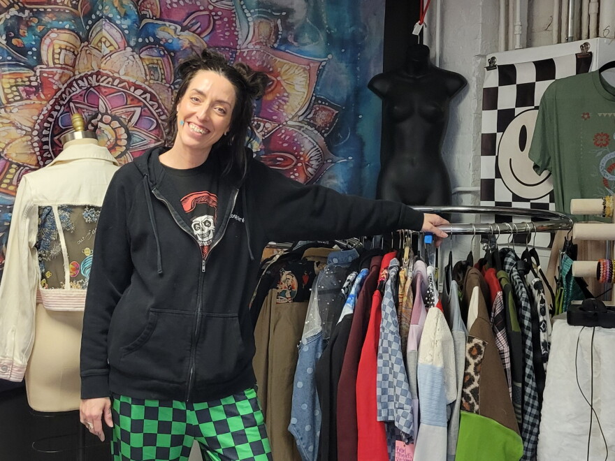 A woman in a black top and green checkered pants smiles next to a rack of clothes
