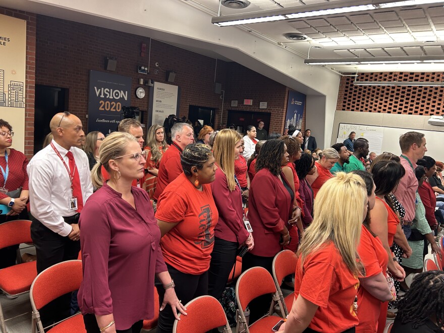 Several dozen adults stand solemnly from chairs in a board room. Most of them are wearing red clothing.