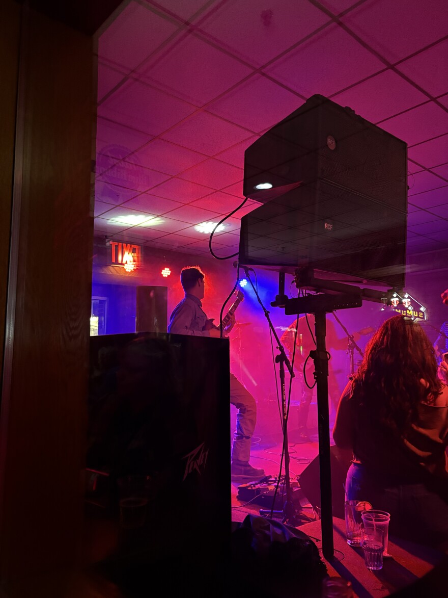 A view from the side of a band performing on a stage bathed in red light. They are seen through a cabinet and speaker on the side of the stage.