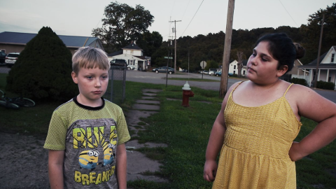 Two children stand on a neighborhood street with a crumbling sidewalk. 
