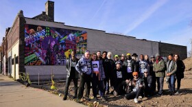 The New State team poses as a group in front of a mural on its building by artist Brad Bernard.