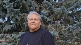 A man dressed in a short-sleeved dark shirt and dark pants poses for a portrait in an outdoor area where you can see a large pine tree behind him. 