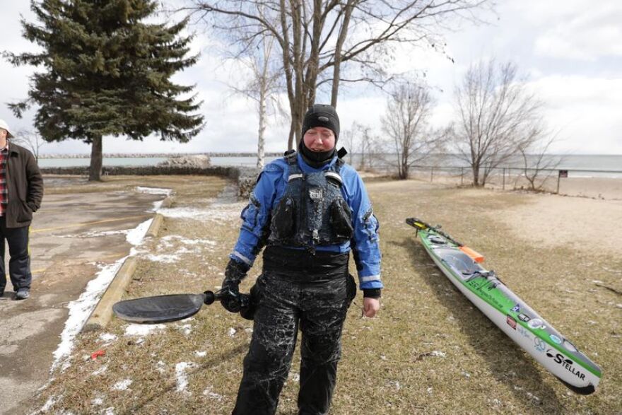 A woman stands in a black wetsuit over a blue jacket, holding a paddle, with a sleek white and green kayak on the ground on a sandy beach, with a lake in the background.