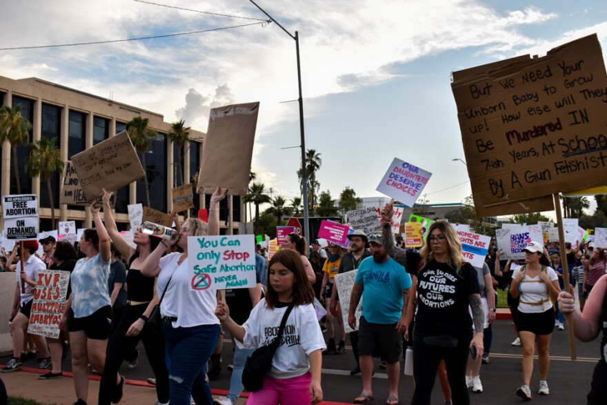 In the crowd of protesters of all ages, a young protester's sign says: You can only stop safe abortion. Never again. Another says: But we need your unborn baby to grow up! How else will they be murdered in 7-ish years, at school, by a gun fetishist?