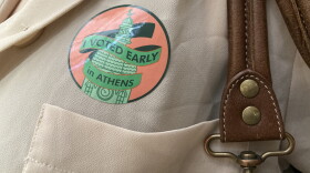 A close-up of an "I voted early in Athens" sticker on someone's beige blouse, next to the brown leather straps of their handbag. 