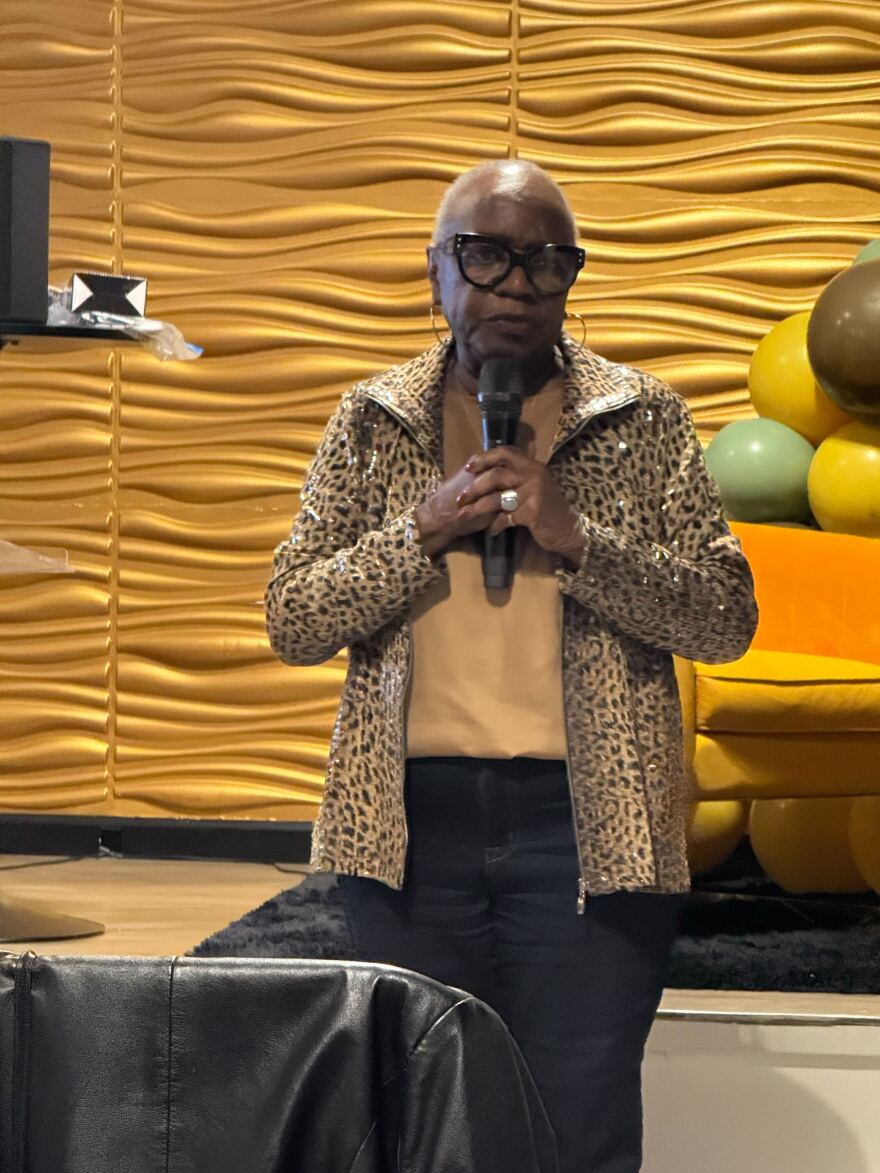 An older Black woman with short hair and a leopard print jacket holds a microphone.