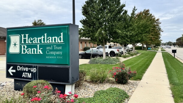 A Heartland Bank and Trust sign directs customers toward the ATM in front of a row of trees.