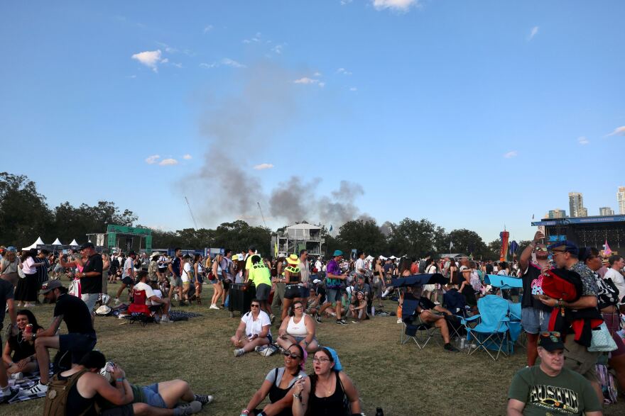 Smoke rises along Austin skyline as seen from Zilker Metropolitan Park, where the third day of Austin City Limits Festival is held on Sunday, Oct. 5, 2025. Festival attendees sit on the grass in the foreground. 