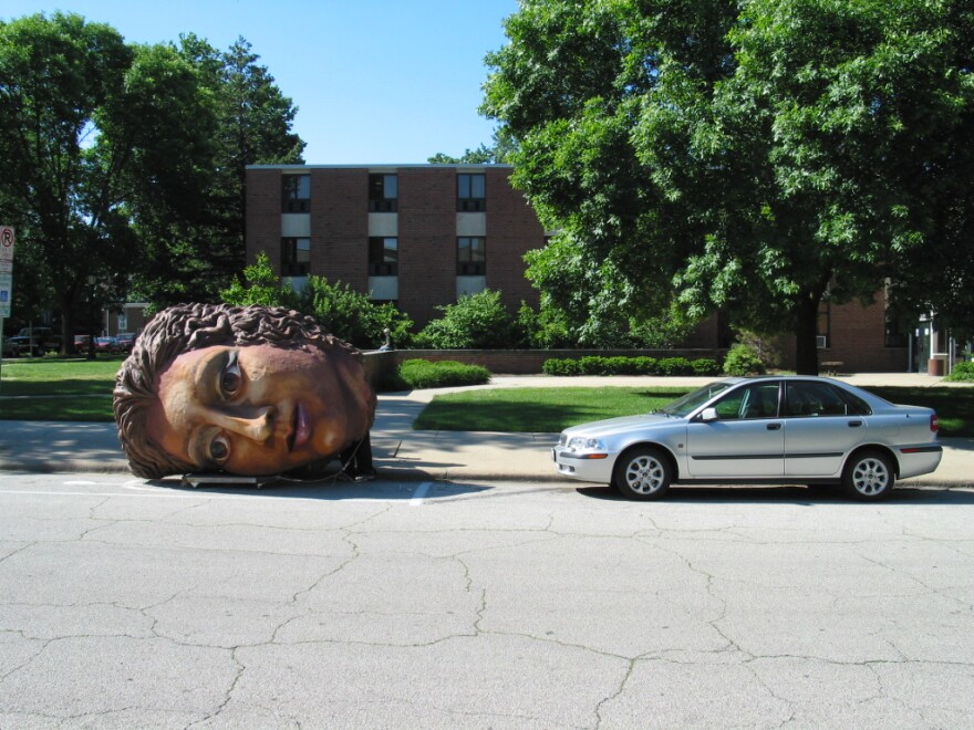 Giant head sits next to car. It is bigger than the car