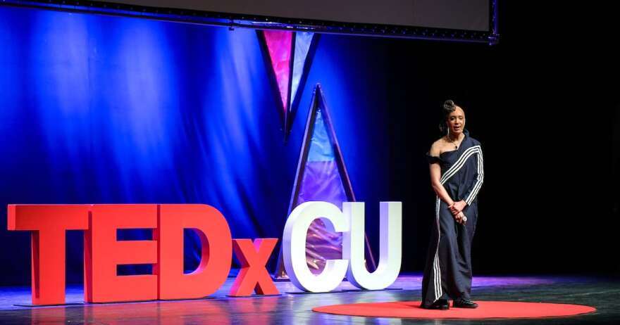 Large letters on stage say TEDxCU. A person stands to the right in a large black gown.