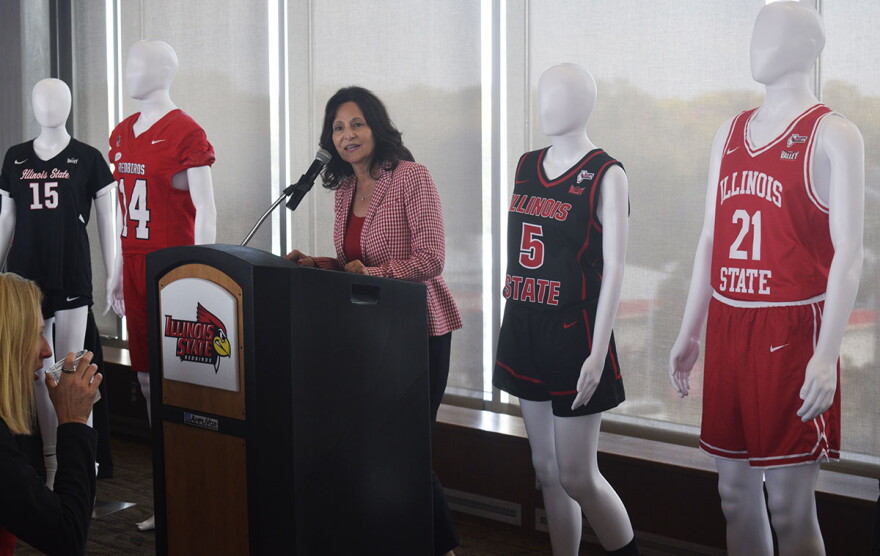 ISU Athletics Director Jeri Beggs speaking at a podium with a series of mannikins at either side. Each displays a sports jersey with 'Illinois State' inscribed on the front. 