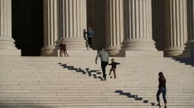 A man holds the hand of a small child as they climb the steps in front of the U.S. Supreme Court Building in Washington, D.C.  A woman follows behind. Their shadows ripple on the steps in front of them.