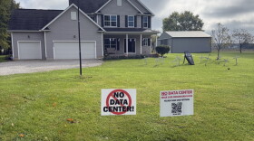 Two signs in front of a large residential home on a rural plot read "No Data Center!" and "No data center near our neighborhoods, please sign our petition," with a QR code.