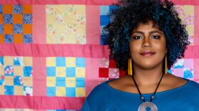 Gragg stands in front of a pink quilt top with yellow and blue squares. She is wearing a large necklace, drop earrings and a blue shirt. Her coily hair has streaks of blue.