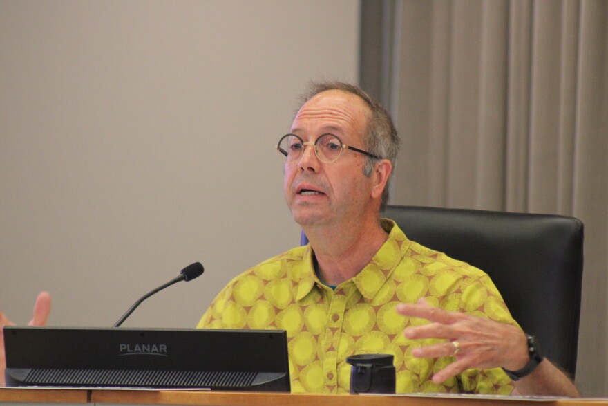 A man in a yellow shirt and glasses sits behind a large desk and computer speaking to someone off camera