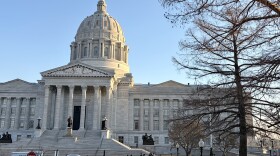 A photo of the exterior of the Missouri State capitol building. Vehicles are parked in front of the domed structure, people walk on the sidewalk. 