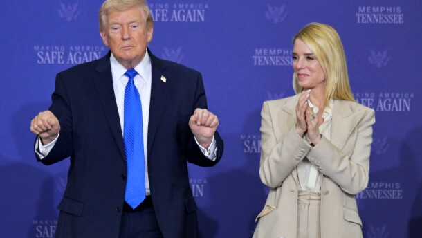 An older white man wearing a dark blur suit stands next to a white middle-aged woman with long blonde hair wearing a beige outfit. She appears to be clapping. 