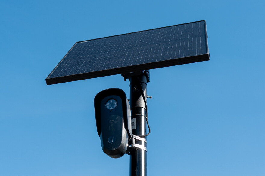 File: Flock license plate reader and camera with solar panel against a blue sky.