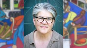 A person in front of a teal background wearing glasses and a blurred painting behind them