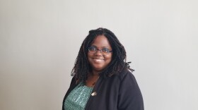 A black woman with glasses stands in front of a white wall smiling