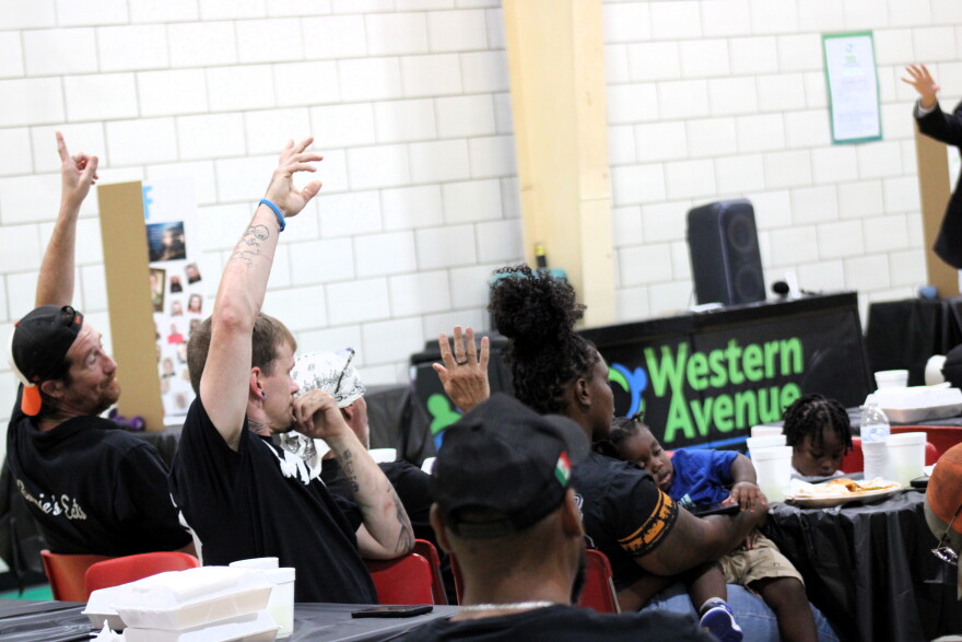 A group of adults sit in tables and chairs, some have their hands raised.