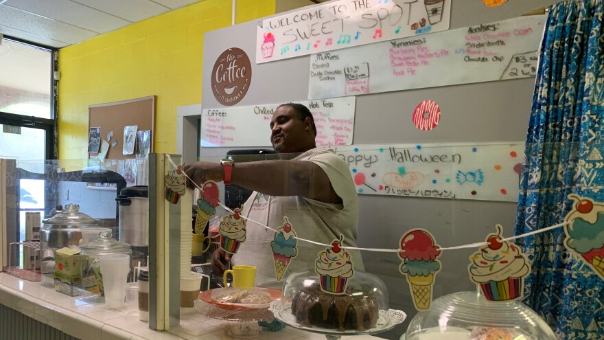 Dabbs making something behind the store counter, next to a sign with the store's name, and a bunch of baked goods in front of him.