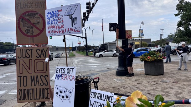 A man holds up several cardboard signs at an intersection as cars drive by. The signs have illustrations of surveillance cameras.