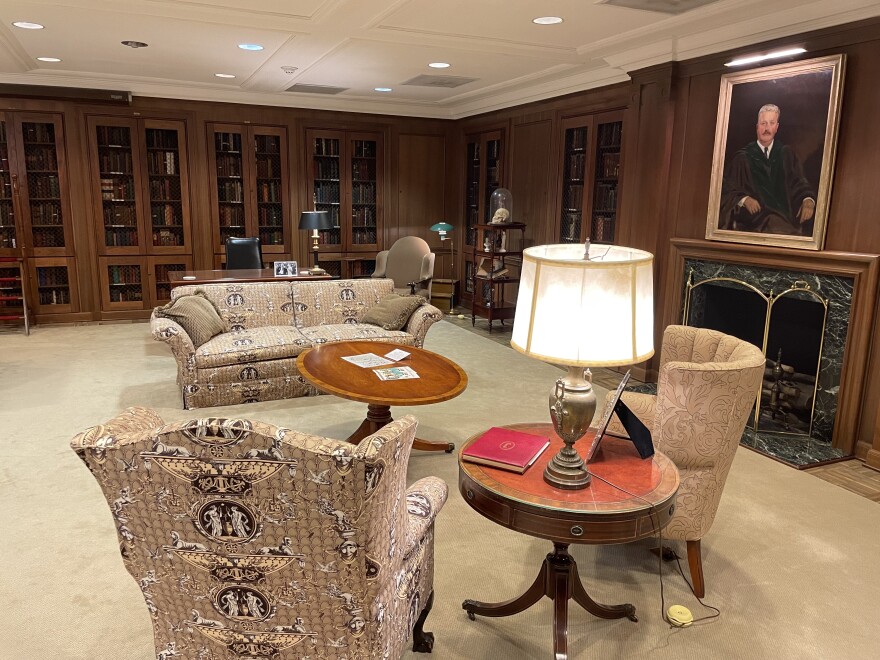 An interior view of a wood-paneled room with a portrait over a fireplace in front of a sofa and armchairs surrounded by bookshelves. 