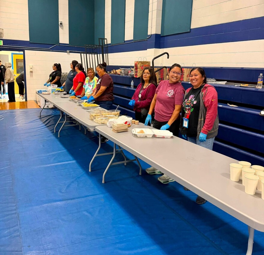 Food placed buffet style on two long tables. Behind the tables are people smiling at the camera or ready to serve food.