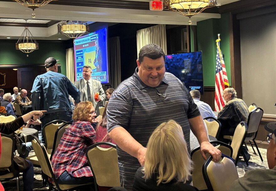 Man standing in a crowded room speaks with people seated at a table