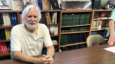 Older man with white hair and beard sits at a table with model ship in front of him. 