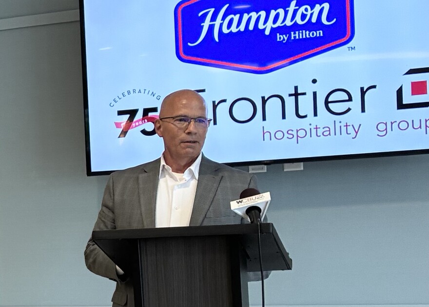 A man in a gray suit and glasses speaks at a podium with microphones. Behind him is a screen displaying "Hampton by Hilton" and "Frontier Hospitality Group.
