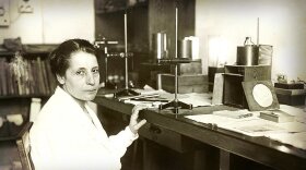 Black-and-white photo of physicist Lise Meitner seated at a laboratory desk, surrounded by scientific instruments and papers, circa 1930.
